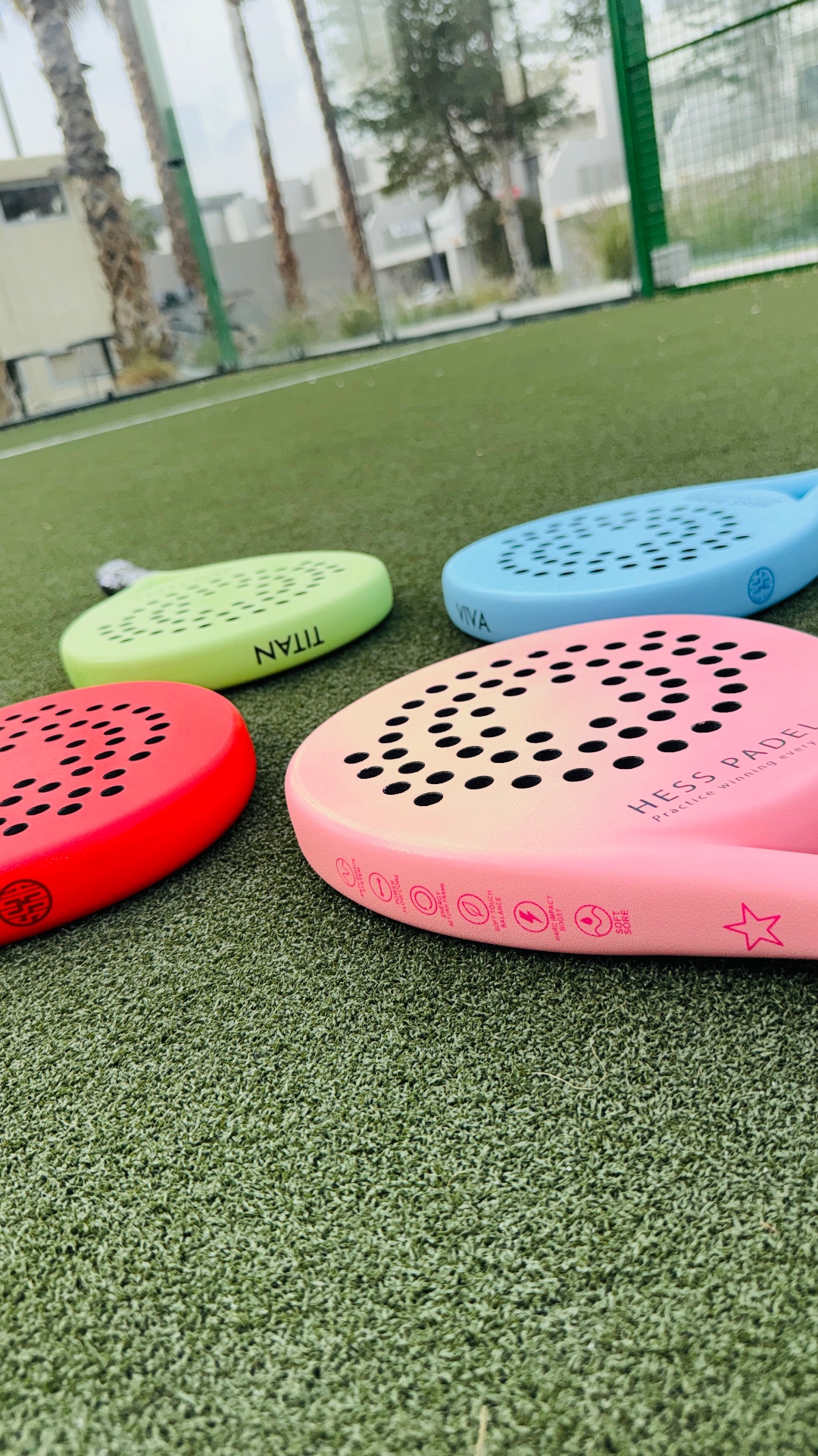 A lineup of HESS PADEL rackets — TITAN lime, VIVA blue, pink, and red — arranged on a Dubai padel court.