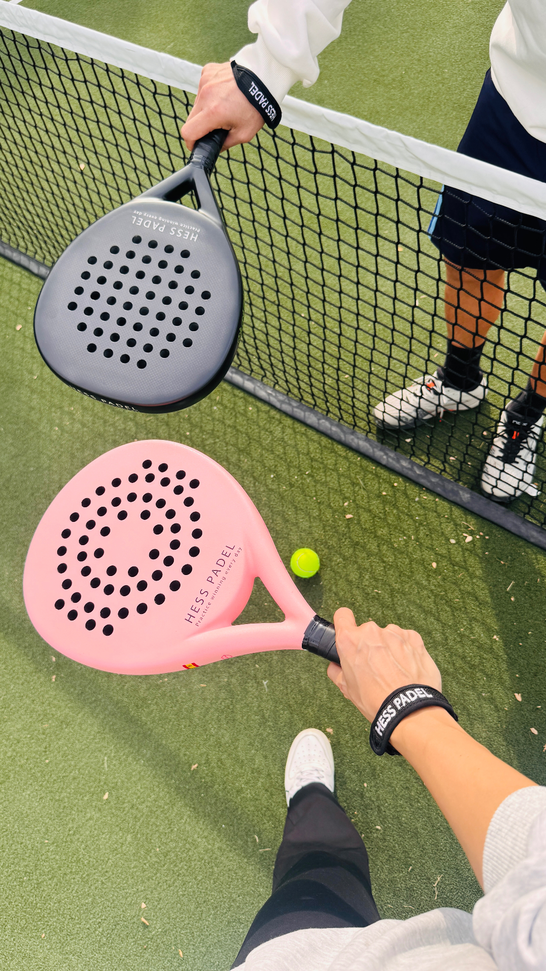 Two players at the net during a padel rally with pink and grey HESS PADEL rackets and HESS PADEL wrist straps.